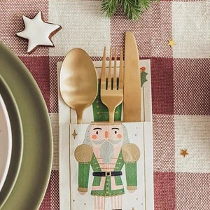 May include: A festive table setting with a gold spoon, fork, and knife. A napkin with a nutcracker design holds the cutlery. The table is set on a red and white checkered tablecloth, with a star-shaped cookie and green plates.