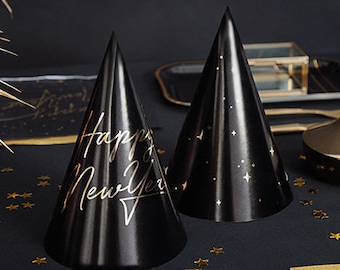 Puede incluir: Dos gorros de fiesta negros con texto dorado «Happy New Year» y diseños de estrellas. Confeti dorado en forma de estrella y platos y servilletas a juego completan el tema festivo de Nochevieja.