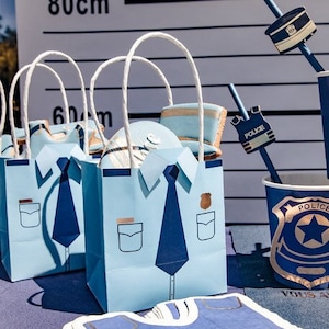 Puede incluir: Tres bolsas de regalo de papel azul y blanco con diseños de temática policial. Las bolsas tienen asas blancas y están decoradas con una camisa azul, corbata y bolsillos. Las bolsas están sobre una mesa con un mantel azul y blanco.