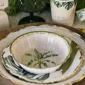 May include: A table setting with a wooden charger, a scalloped plate with a gold rim, and a bowl with a palm tree design. A green glass, a paper cup, and a small glass bottle are also present. The setting has a tropical theme.