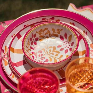 Könnte beinhalten: Eine lebendige Tischdekoration mit rosa und weiß gemusterten Tellern, einer kleinen Schüssel mit floralen Designs und zwei dekorativen Gläsern. Die Teller haben einen goldenen Rand und sind auf einer rosa gestreiften Tischdecke angeordnet. Die Gläser sind orange.