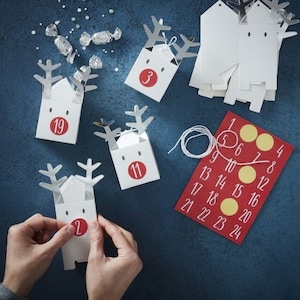 May include: White reindeer-shaped advent calendar boxes with silver antlers and red numbered circles. A red calendar card with numbers 1-24 and yellow circles is also present. The image is on a dark blue surface.