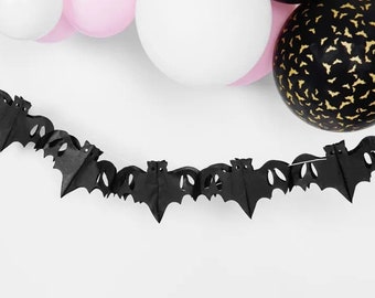 May include: A black paper bat garland is strung across the image, with a series of bat cutouts. Balloons in white, pink, and black with gold bat silhouettes are in the background. This festive decoration is perfect for Halloween.