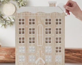 May include: Wooden advent calendar in the shape of a house. The house has 24 numbered windows, with a white snow-covered roof and trim. A hand is peeling off a white sticker. The house sits on a brown base.