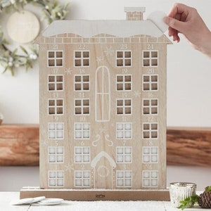 Könnte beinhalten: Adventskalender aus Holz in Form eines Hauses. Das Haus hat 24 nummerierte Fenster mit weißem, schneebedecktem Dach und Zierleisten. Eine Hand löst einen weißen Aufkleber ab. Das Haus steht auf einem braunen Sockel.