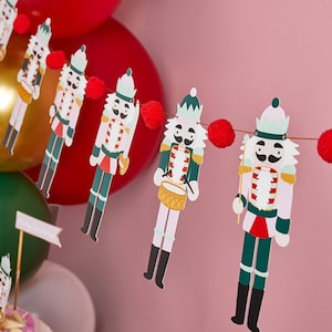 Puede incluir: Una guirnalda festiva con una cadena de figuras de cascanueces con pompones rojos entre las figuras. Los cascanueces están vestidos con atuendos tradicionales con acentos verdes, rojos y blancos.