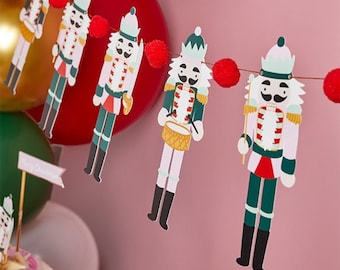 May include: A festive garland featuring a string of nutcracker figures with red pom-poms in between. The nutcrackers are dressed in traditional attire with green, red, and white accents.