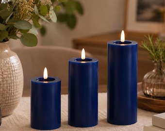 Könnte beinhalten: Drei zylindrische blaue Stumpenkerzen in unterschiedlichen Höhen, jede mit einer kleinen, leuchtenden Flamme an der Spitze. Die Kerzen haben eine glatte, einfarbig blaue Wachsoberfläche und sind in einer Reihe auf einem strukturierten Stoffläufer angeordnet.