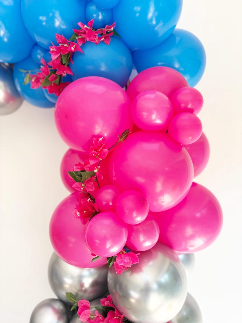 May include: A vibrant balloon arrangement featuring blue, pink, and silver balloons. Pink flowers are interspersed throughout the arrangement. The balloons vary in size, creating a dynamic and festive display.