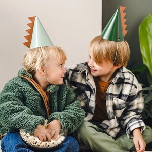 May include: Two children wearing green and brown dinosaur party hats are sitting on a white rug. The child on the left is holding a bowl of popcorn.