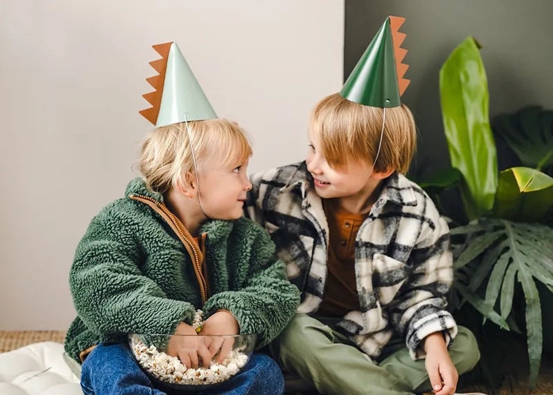 May include: Two children wearing green and brown dinosaur party hats are sitting on a white rug. The child on the left is holding a bowl of popcorn.