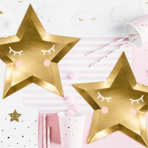 May include: Two gold star-shaped paper plates with a pink and white sleeping face design. The plates are on a pink and white striped background. There are also pink and white cups and straws in the image.
