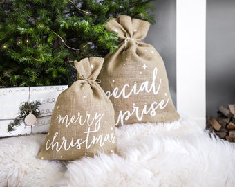Puede incluir: Dos bolsas de regalo de arpillera con texto blanco "merry christmas" y "special prize". Las bolsas están decoradas con pequeñas formas de estrella blanca. Las bolsas están sobre una superficie blanca y esponjosa.