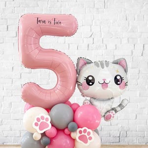 May include: A pink number 5 balloon with the text "Tara is Five" is the centerpiece of a balloon arrangement. A cat-shaped balloon and a cluster of pink, grey, and white balloons with paw prints complete the festive display.