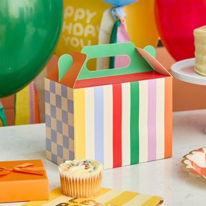 May include: A rectangular cardboard party favour box with a handle, featuring a multicoloured vertical striped pattern on the front and a light purple and beige checkered pattern on the side, with orange and green accents on the top flaps.