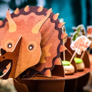 May include: A detailed cardboard Triceratops dinosaur model in brown and orange. The model has a frill, horns, and a detailed body. The dinosaur is part of a themed display with food items.