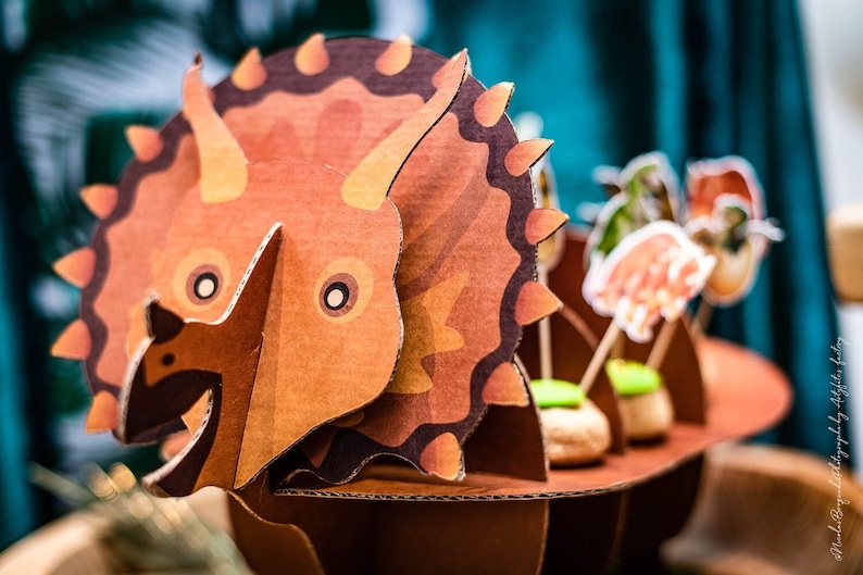May include: A detailed cardboard Triceratops dinosaur model in brown and orange. The model has a frill, horns, and a detailed body. The dinosaur is part of a themed display with food items.