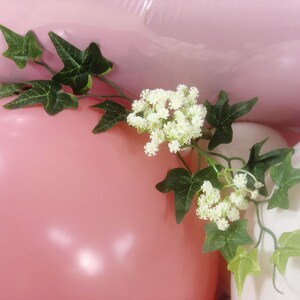 May include: Pink and white balloons with green ivy and white flowers. The balloons are tied together and the ivy and flowers are draped over the balloons.