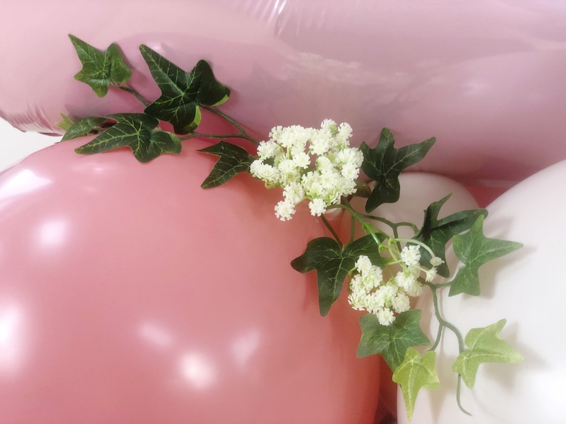 May include: Pink and white balloons with green ivy and white flowers. The balloons are tied together and the ivy and flowers are draped over the balloons.