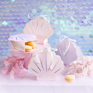 May include: Four pink and purple seashell-shaped gift boxes with white ribbon. The boxes are decorated with a pearl-like shimmer. Two of the boxes are filled with small, round, yellow cookies.