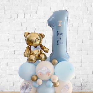 May include: A gold teddy bear balloon with a blue bow tie sits atop a cluster of blue and white balloons. A large blue number one balloon with the text "Tara is One" is also present. The balloons are decorated with baby-themed graphics.