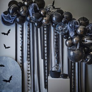 May include: A Halloween-themed party decoration featuring a black balloon arch with bat cutouts and faux spiderwebs. Black and white striped fabric hangs behind the balloons. A cauldron and bowl sit on a white pedestal, with bat decorations on the wall.