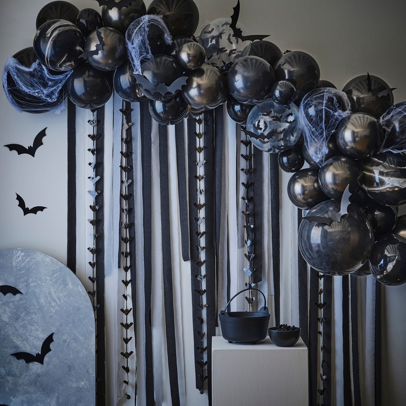 May include: A Halloween-themed party decoration featuring a black balloon arch with bat cutouts and faux spiderwebs. Black and white striped fabric hangs behind the balloons. A cauldron and bowl sit on a white pedestal, with bat decorations on the wall.