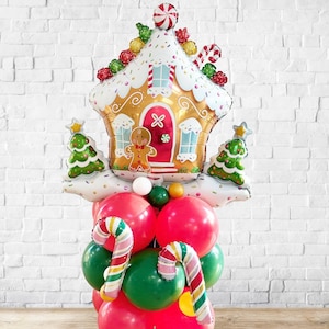 May include: A festive Christmas balloon arrangement featuring a gingerbread house, candy canes, and Christmas trees. The gingerbread house is gold with white frosting and red accents. The base is composed of red and green balloons.