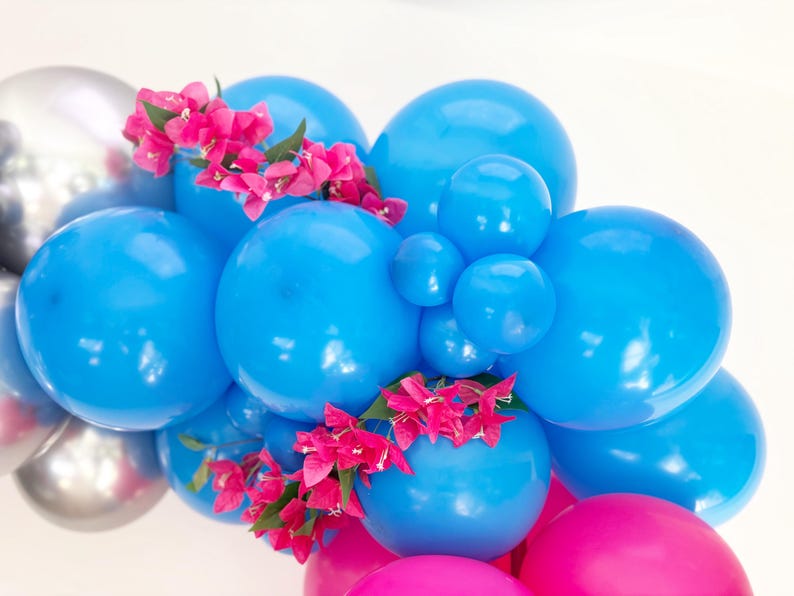 May include: A cluster of blue, silver, and pink balloons, adorned with bright pink flowers. The balloons vary in size, creating a dynamic and festive arrangement. Ideal for party decorations.