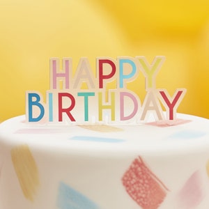 May include: A white cake with colorful brushstrokes on the frosting. A multi-colored "Happy Birthday" topper sits on top of the cake.
