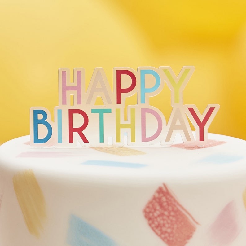 May include: A white cake with colorful brushstrokes on the frosting. A multi-colored "Happy Birthday" topper sits on top of the cake.