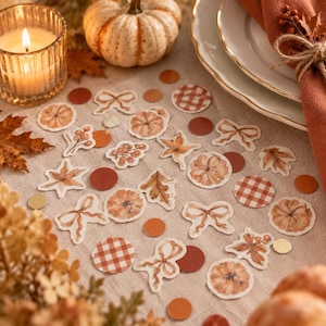 Könnte beinhalten: Eine Sammlung von herbstlichem Papierkonfetti mit Illustrationen von Kürbissen, Schleifen, Blättern, Beeren und orangefarbenen Karomustern. Das Set enthält verschiedene kreisförmige Formen in einfarbigem Orange und Braun auf einer neutralen Stoffoberfläche.