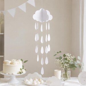 May include: A white cloud-shaped mobile with hanging teardrop decorations hangs above a table set for a baby shower. The table features a cake, cupcakes, and floral arrangements. A sign says "Oh baby".