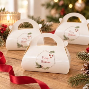 May include: Three white cardboard gift boxes with handles, each featuring a printed geometric frame, green pine branch illustrations, red berries, and the text Merry Christmas in red script lettering on the front panel.