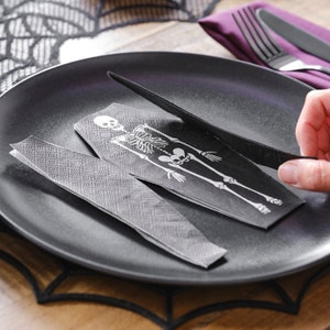 May include: Black and white skeleton printed on a black napkin, folded into a coffin shape, on a black plate. The napkin is being cut with a black knife.