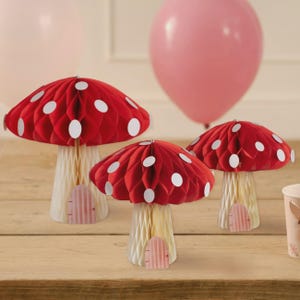 May include: Three red mushroom decorations with white polka dots and cream-coloured stems. Each mushroom has a small pink door. Two pink balloons are in the background. The mushrooms are on a wooden surface.