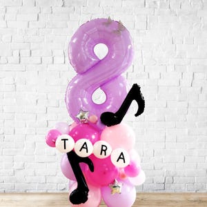 Könnte beinhalten: Eine Ballonanordnung mit einer großen, lilafarbenen Acht, darunter kleinere rosa und weiße Ballons. Schwarze Noten und der Name "TARA" sind ebenfalls enthalten, mit sternförmigen Akzenten. Die Ballons sind in einer Säule angeordnet.