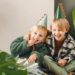 May include: Two children wearing green and brown dinosaur party hats are sitting together. One child is eating popcorn from a bowl while the other child is laughing. The children are surrounded by green plants.