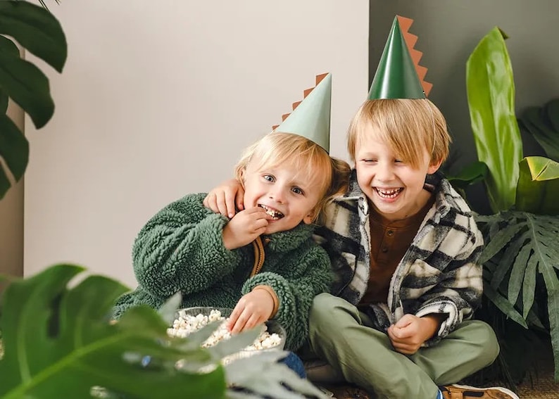 May include: Two children wearing green and brown dinosaur party hats are sitting together. One child is eating popcorn from a bowl while the other child is laughing. The children are surrounded by green plants.