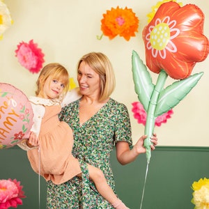 May include: A woman and a young girl are holding pink and red flower-shaped balloons. The balloons have the text "Dla Kochanej MAMY" printed on them. The woman is wearing a green floral dress and the girl is wearing a peach dress. They are standing in front of a wall decorated with paper flowers.