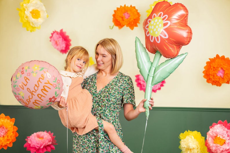 May include: A woman and a young girl are holding pink and red flower-shaped balloons. The balloons have the text "Dla Kochanej MAMY" printed on them. The woman is wearing a green floral dress and the girl is wearing a peach dress. They are standing in front of a wall decorated with paper flowers.