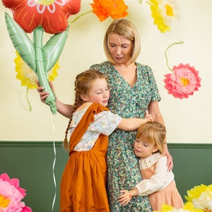 May include: A woman and two girls are standing in front of a wall decorated with paper flowers. The woman is holding a large red and green flower-shaped balloon. The girls are hugging each other.