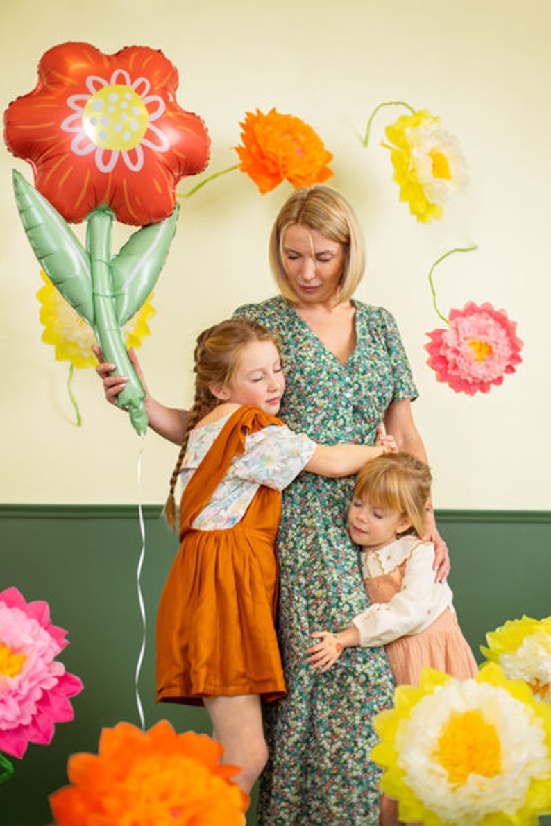 May include: A woman and two girls are standing in front of a wall decorated with paper flowers. The woman is holding a large red and green flower-shaped balloon. The girls are hugging each other.