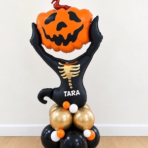 May include: A balloon sculpture featuring a black cat body with a gold skeleton print and the name TARA in white text. The cat holds an orange jack-o'-lantern balloon above its head. The base consists of stacked black, gold, orange, and white balloons.