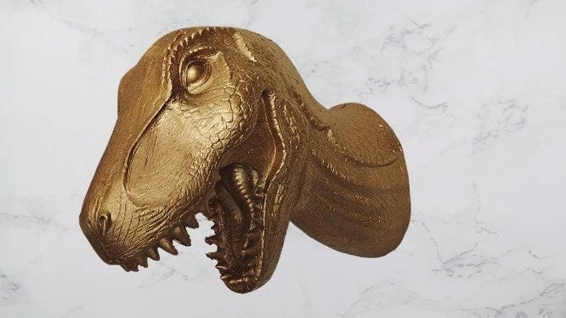 May include: A gold-coloured Tyrannosaurus Rex head sculpture with detailed texture and open mouth, showcasing sharp teeth. The sculpture is mounted on a wall, adding a touch of prehistoric decor.