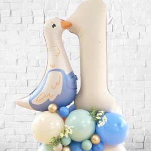 May include: A celebratory balloon display for a first birthday. Includes a large number one balloon and a goose-shaped balloon. The base is made of blue, green, and cream-coloured balloons, with gold spheres and small white flowers as accents.