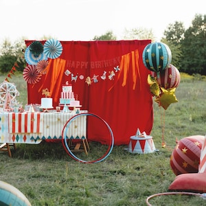 Op de afbeelding: Een feestelijke outdoor verjaardagsopstelling met een circusthema. Inclusief een tafel met een taart, reuzenrad en decoraties, een rode gordijnachtergrond, ballonnen en een gestreepte tent. De sc&egrave;ne speelt zich af in een grasveld.