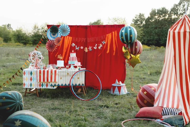 Op de afbeelding: Een feestelijke outdoor verjaardagsopstelling met een circusthema. Inclusief een tafel met een taart, reuzenrad en decoraties, een rode gordijnachtergrond, ballonnen en een gestreepte tent. De sc&egrave;ne speelt zich af in een grasveld.