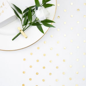 Puede incluir: Un plato blanco con un borde dorado, una pequeña caja blanca con el texto "Gracias" en letras doradas, una sola flor blanca con hojas verdes y una pinza de ropa dorada. El plato está sobre una superficie blanca con confeti dorado.