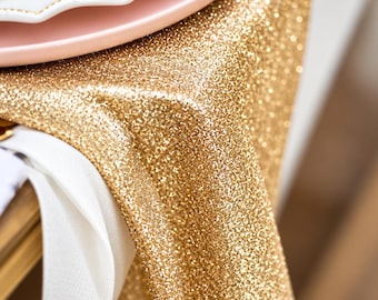Puede incluir: Un camino de mesa con purpurina dorada drapeado sobre una superficie, con una textura densa y brillante. Un plato rosa pálido con un borde festoneado blanco y una servilleta blanca están colocados en el lado izquierdo de la tela dorada.